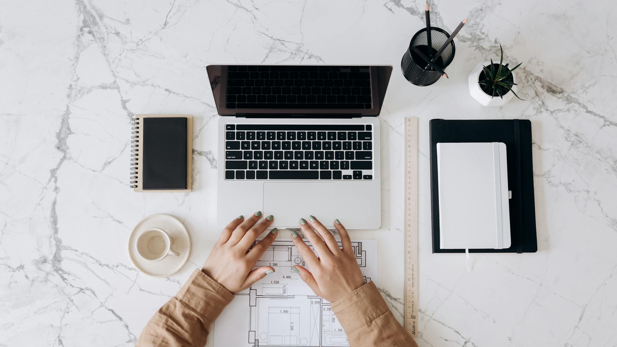 top view of a stylish home office desk with a laptop planner and coffee cup showing hands on a blueprint. 6894103 scaled